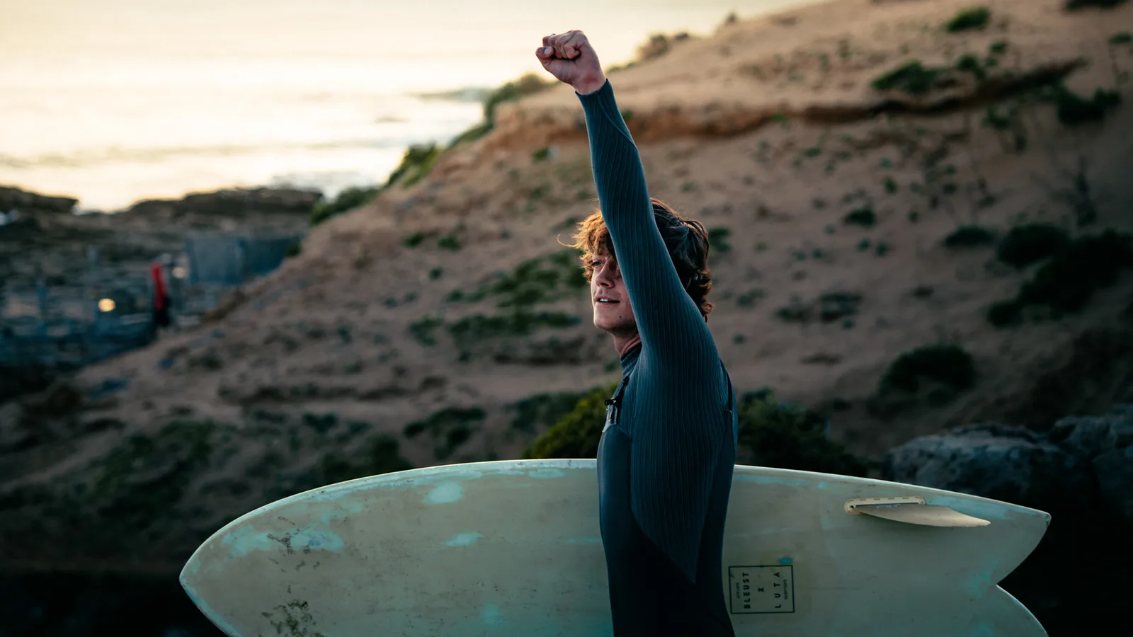 Surfer holding his board and pumping a fist above a rugged coastal cliff in Essaouira, Morocco
