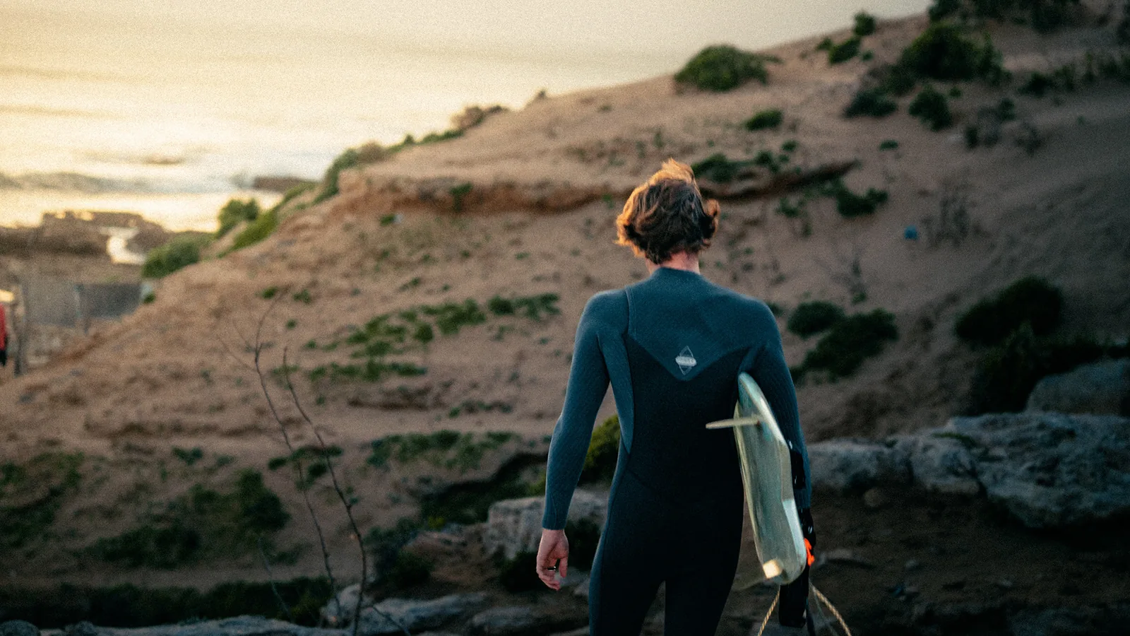 Surfer walking inland along a sandy dune trail with his board at sunset in Essaouira, Morocco