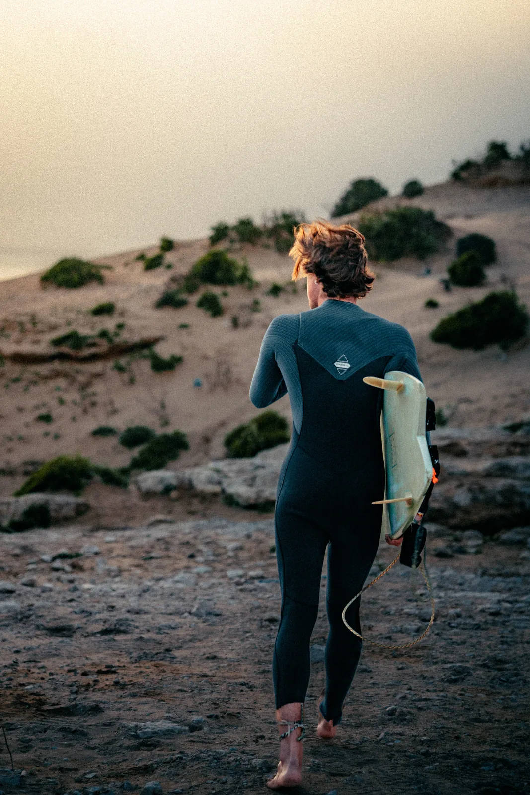 Surfer strolling across a dune path toward the shoreline at dusk in Essaouira, Morocco