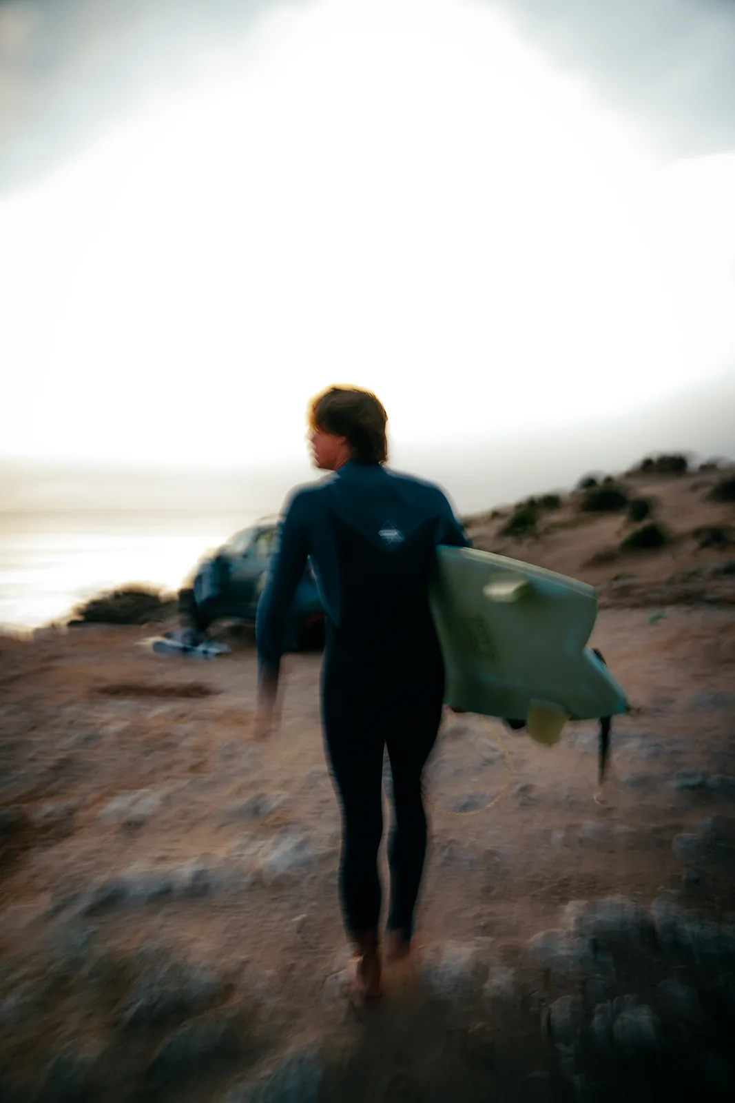 Soft-focus surfer walking a dune path bathed in warm evening light in Essaouira, Morocco