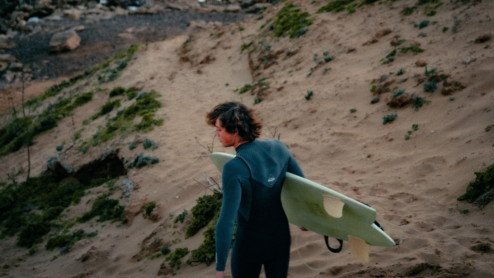 Surfer glancing back while climbing a sandy dune path with his board in Essaouira, Morocco