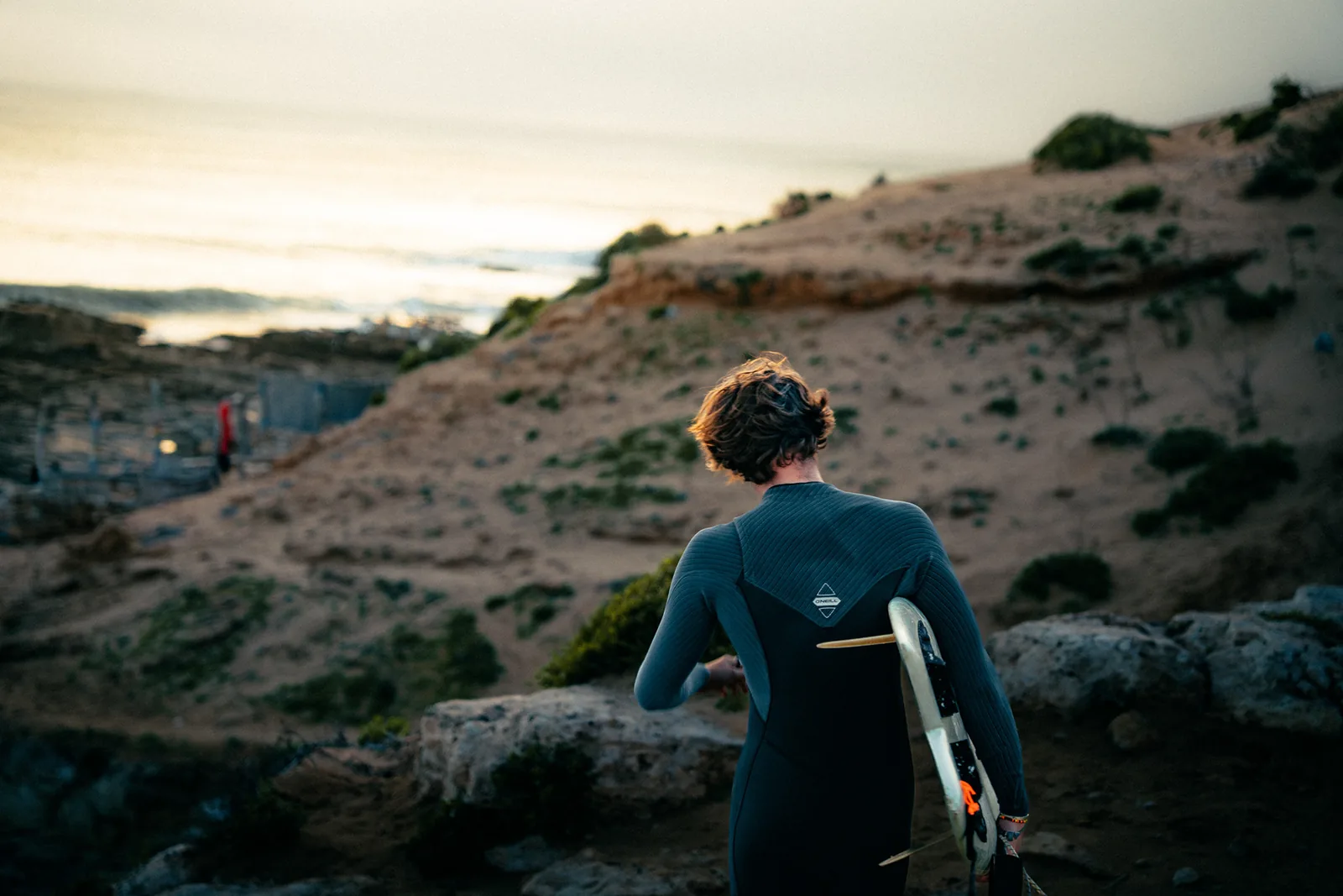 Surfer heading toward a rocky outcrop at dusk, board tucked under his arm, in Essaouira, Morocco