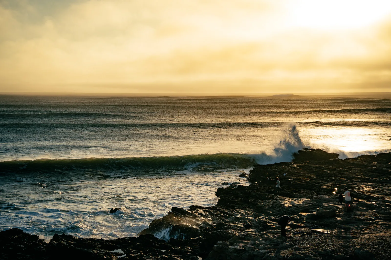 Onlookers watching waves burst over dark rocks as the sun sets in Essaouira, Morocco