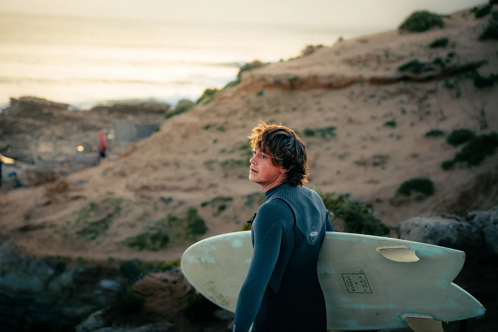 Golden-hour light catches a surfer on a coastal bluff, board in hand, facing the sea in Essaouira