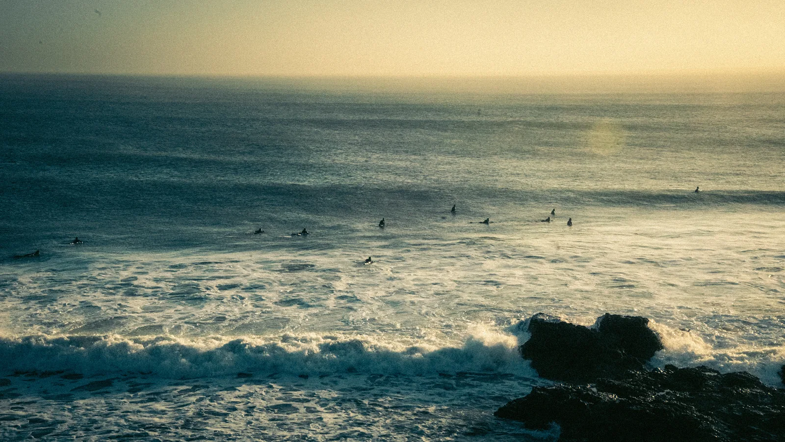 Surfers wait in a golden lineup as waves break against dark rocks in Essaouira, Morocco