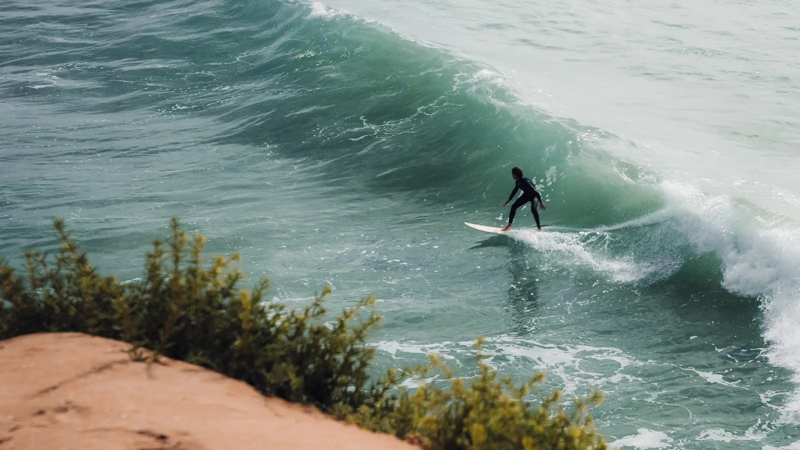Surfer crouched low while riding a turquoise wave past a rugged cliff in Essaouira, Morocco
