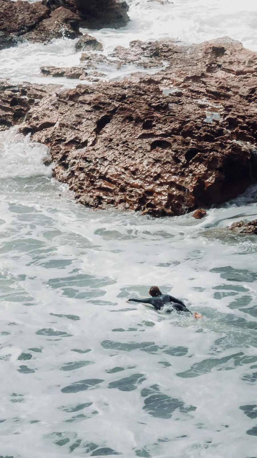 Surfer paddling past a jagged rock formation through foaming water in Essaouira, Morocco