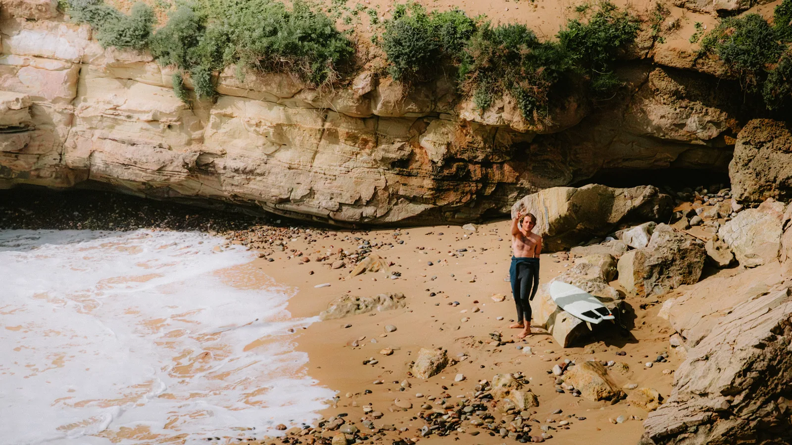 Surfer standing beside a board on a remote cove beach beneath sandstone cliffs in Essaouira, Morocco