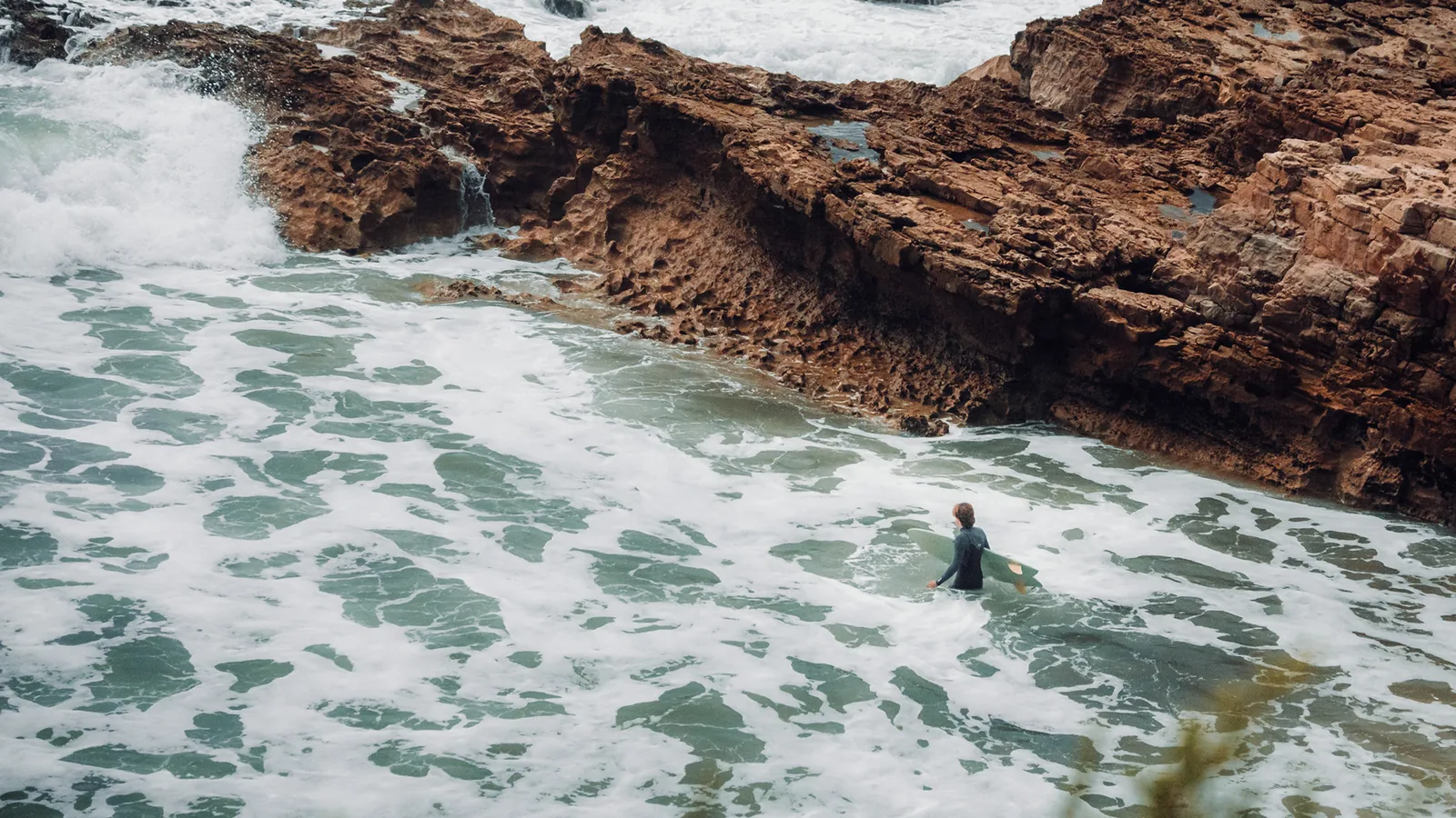 Surfer wading chest-deep near a jagged rocky outcrop in Essaouira, Morocco