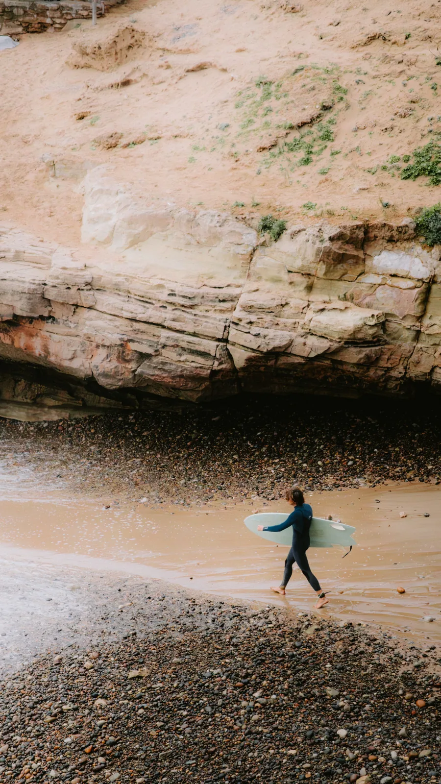 Surfer carrying his board along a wet cove beach beneath overhanging cliffs in Essaouira, Morocco