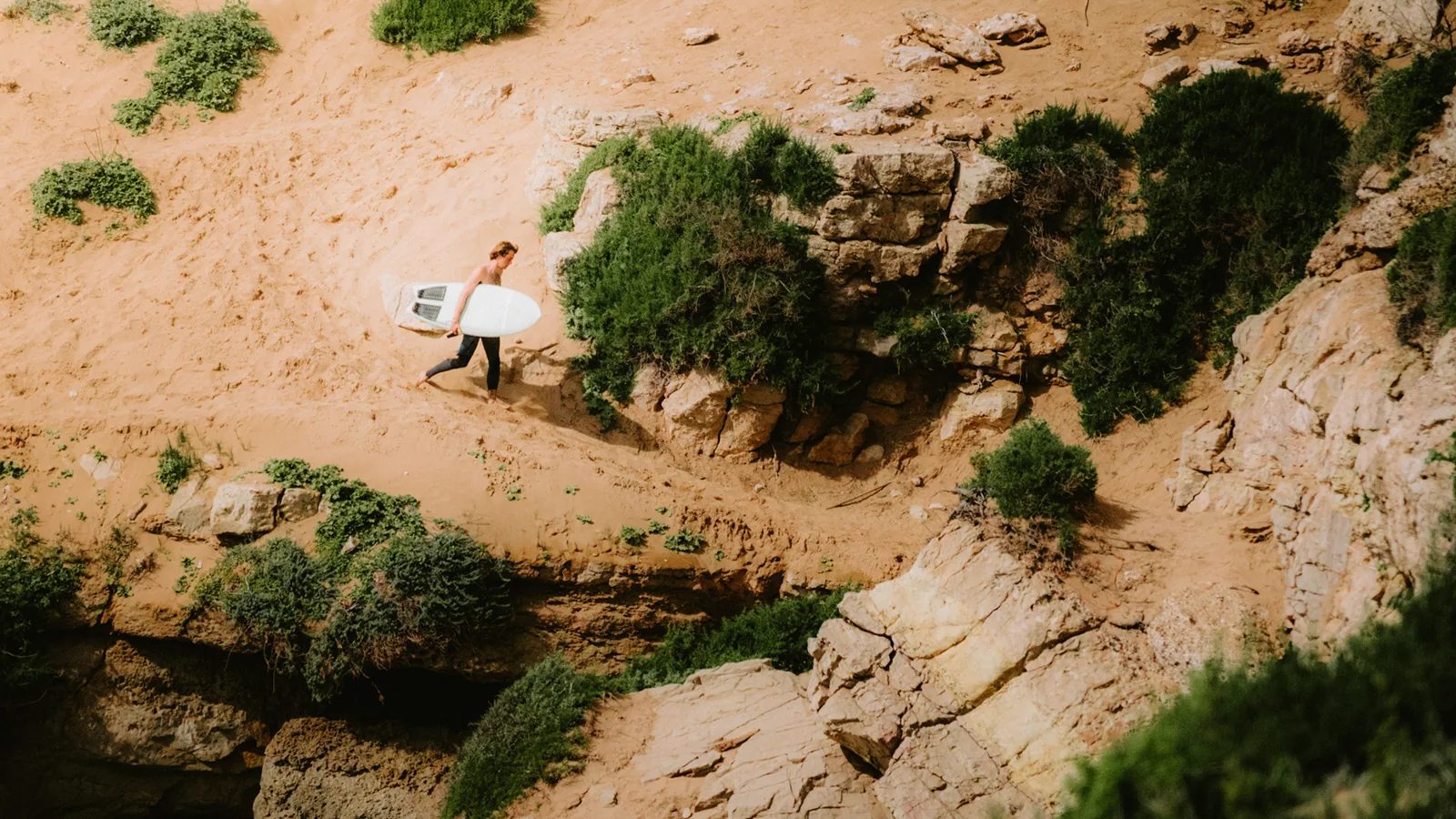 Seen from above, a surfer crosses a sandy clifftop carrying a board at Essaouira, Morocco