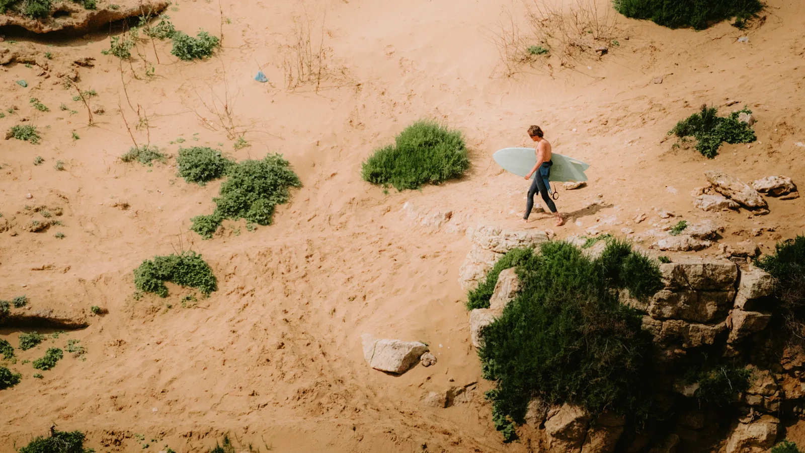 An aerial view shows a surfer walking across dunes toward the cliff edge at Essaouira