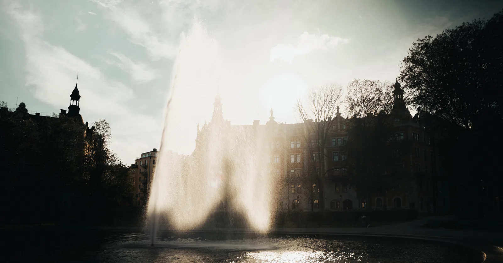 Backlit fountain mist drifting over a tranquil city park pond in Sweden