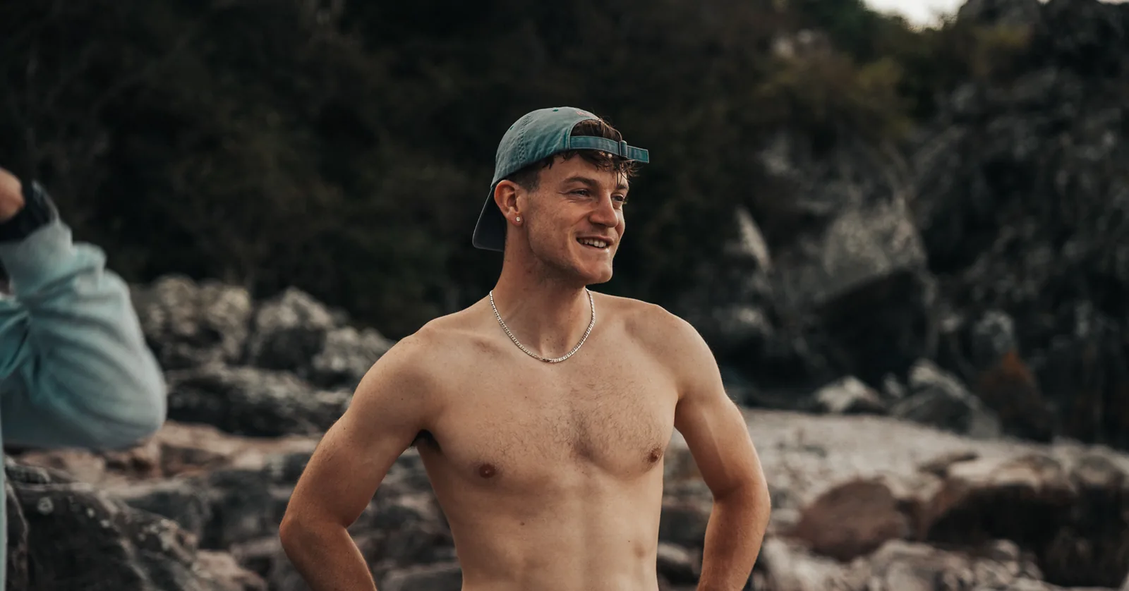 Shirtless man smiling with hands on hips along a rocky shoreline in Sweden