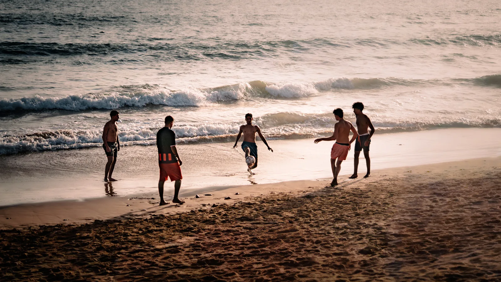 Young men play beach football as waves roll in at sunset near Taghazout, Morocco