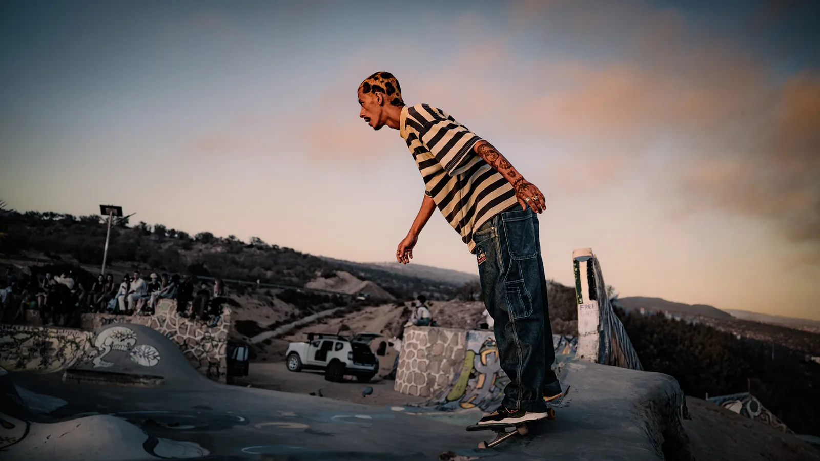 A skater balances atop a concrete bowl overlooking parked cars at sunset, Taghazout