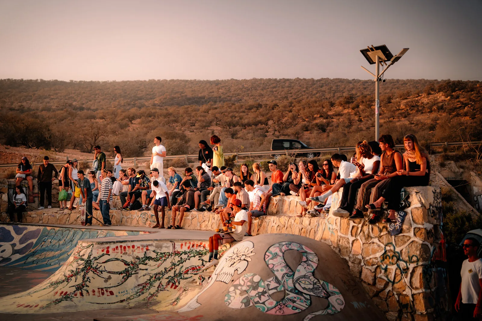 A crowd gathers along the rim of a skate bowl to watch riders at sunset, Taghazout