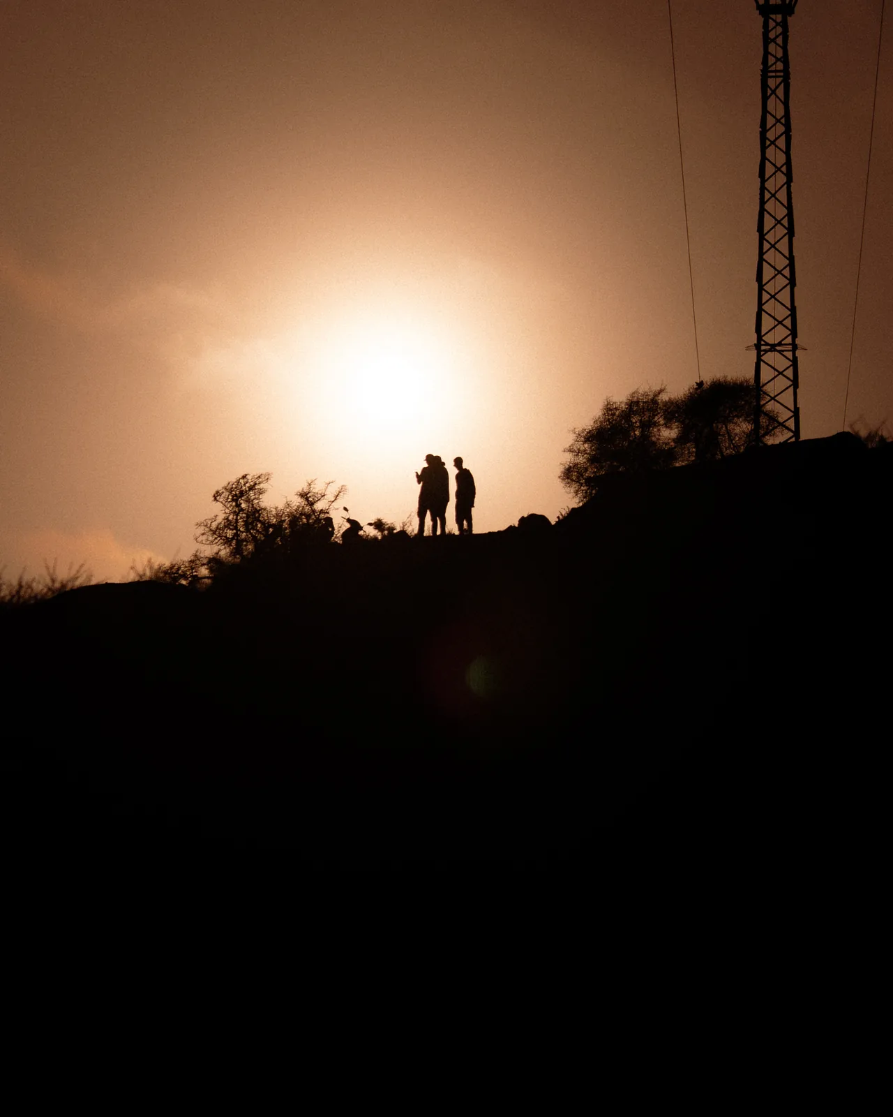 Two silhouetted figures stand on a hilltop beneath a lighting tower at sunset, Taghazout