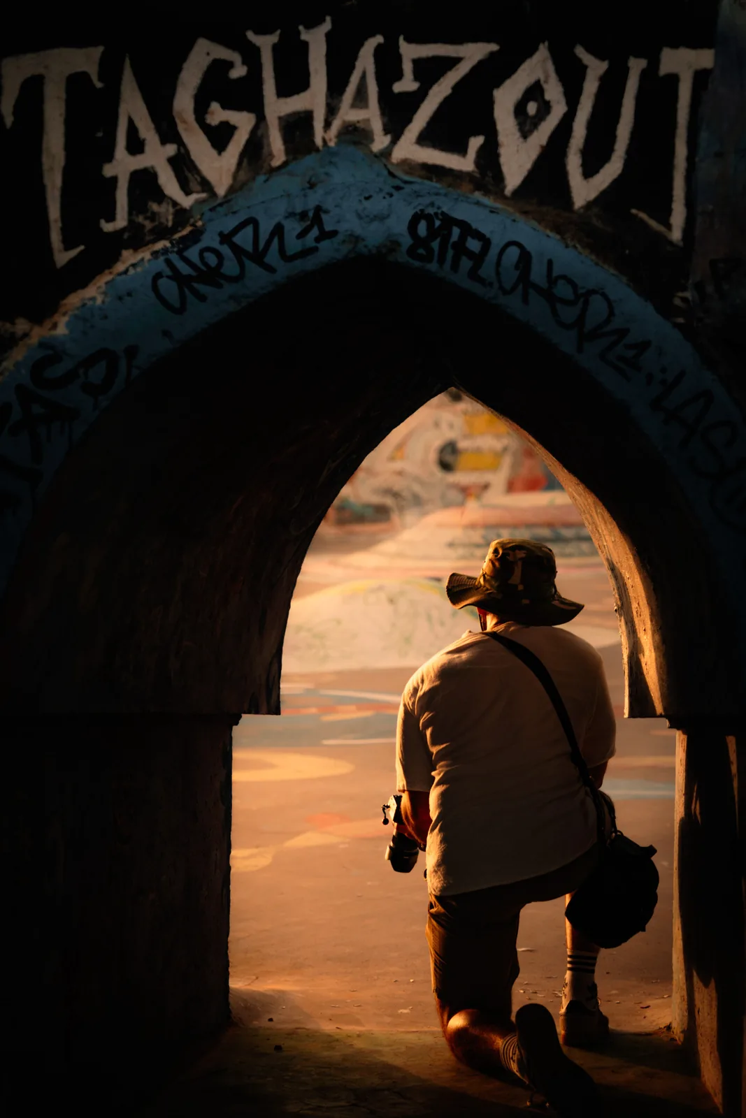A photographer crouches beneath a graffiti-tagged archway reading Taghazout, Morocco