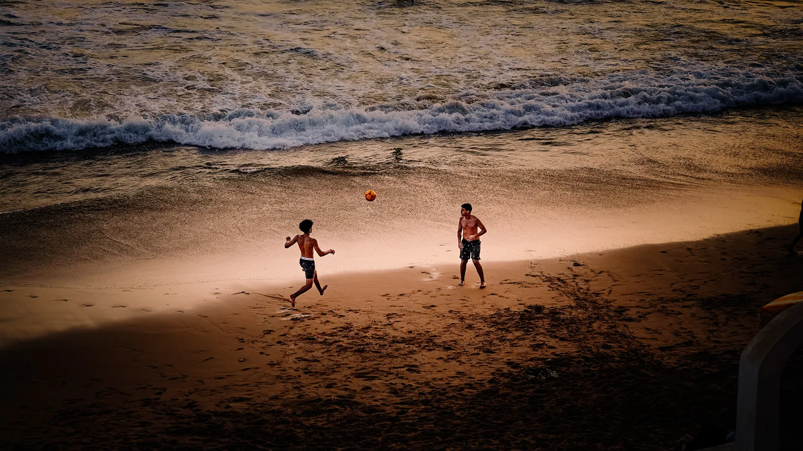 Two boys chasing a ball across the sand as the sun dips low in Taghazout, Morocco